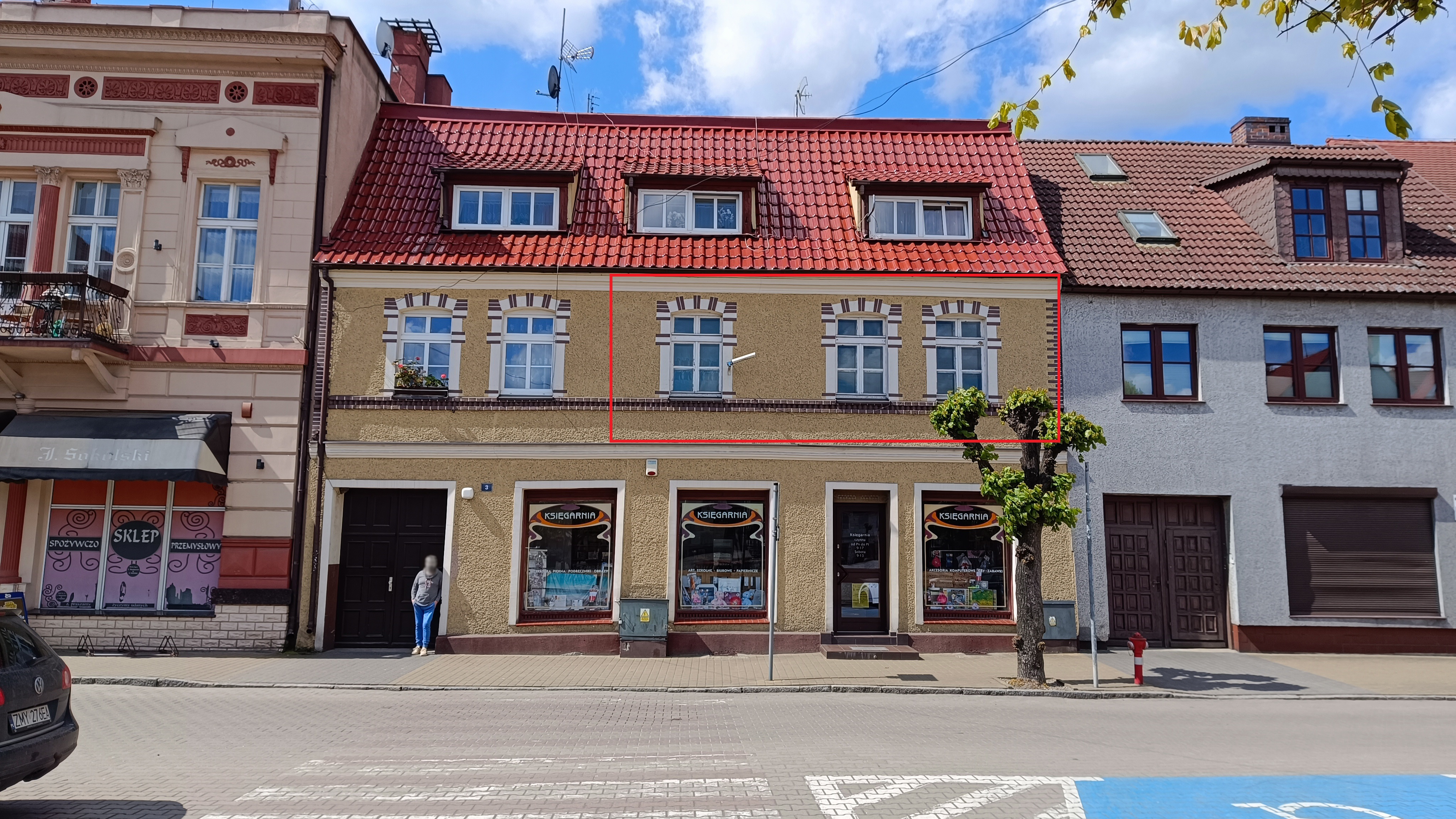 Budynek przy ul. Kościuszki w Trzcińsku-Zdroju. Na parterze znajdują się dwa lokale usługowe, w tym księgarnia. Pierwsze piętro, oznaczone czerwoną ramką, to mieszkanie nr 3 o powierzchni 65,31 m², wystawione na sprzedaż przez Gminę Trzcińsko-Zdrój. Budynek ma jasnobeżową elewację z dekoracyjnymi ceglanymi wstawkami wokół okien i dach pokryty czerwonymi dachówkami. Na chodniku przed budynkiem znajduje się mężczyzna oparty o drzwi wejściowe.